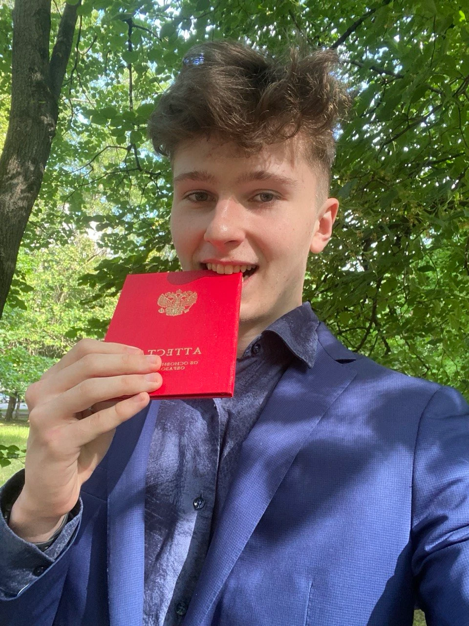 Dmitrii biting his red diploma with honors in a summer park